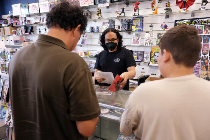 A store manager, Nicholas Ignacio, wearing a mask, serves two customers from behind the counter.